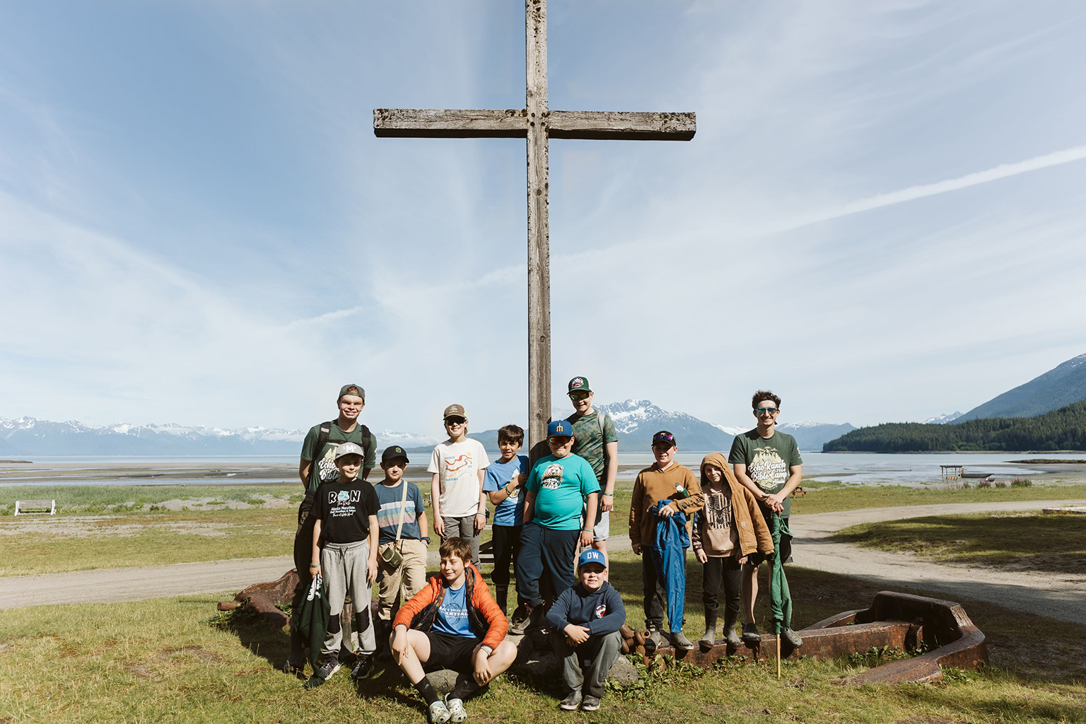 Group photo of Evan and Echo Ranch campers standing in front of a rustic wooden cross with majestic Alaskan mountains and lake in the background.